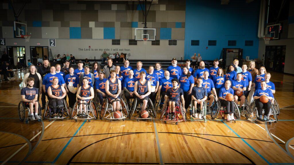 A group photo in the middle of the basketball court of all of the participants and volunteers for the 2026 Wheelchair Basketball event